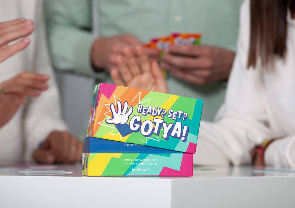 Two colorful "Ready? Set? GOTYA!" game boxes on a white surface, with blurred people playing in the background