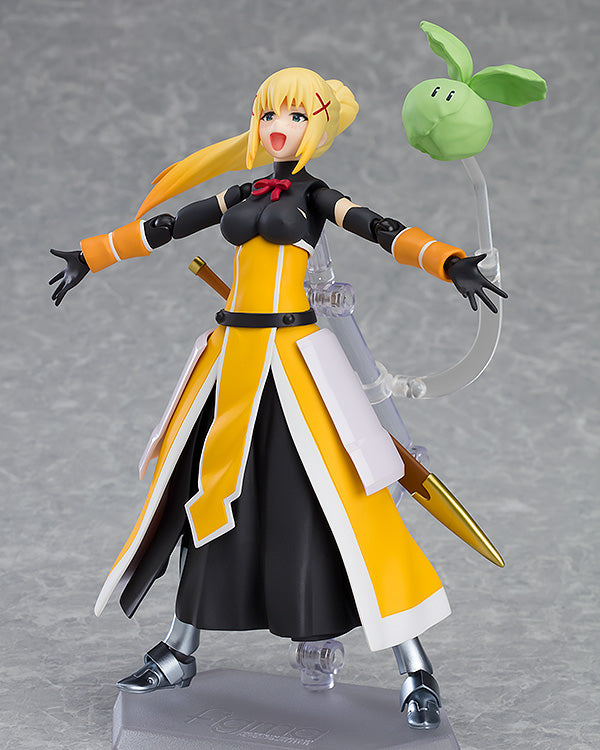 A Figma Darkness figure from KonoSuba stands dynamically with outstretched arms, an open-mouthed smile, and her signature yellow and white outfit, with a floating green cabbage beside her