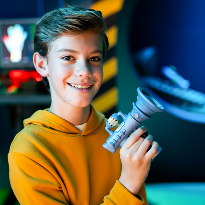A cheerful boy with braces in a yellow hoodie holds a light blue Moose Despicable Me 4 Mini Fart Blaster with a yellow and black caution-striped tank