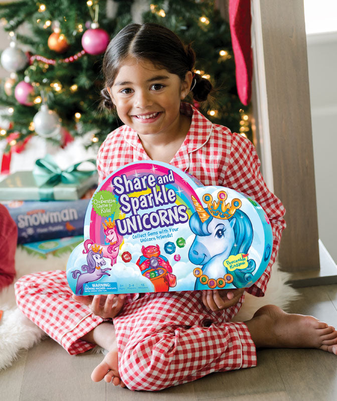 Child in pajamas holding &quot;Share and Sparkle Unicorns&quot; Peaceable Kingdom game by Christmas tree