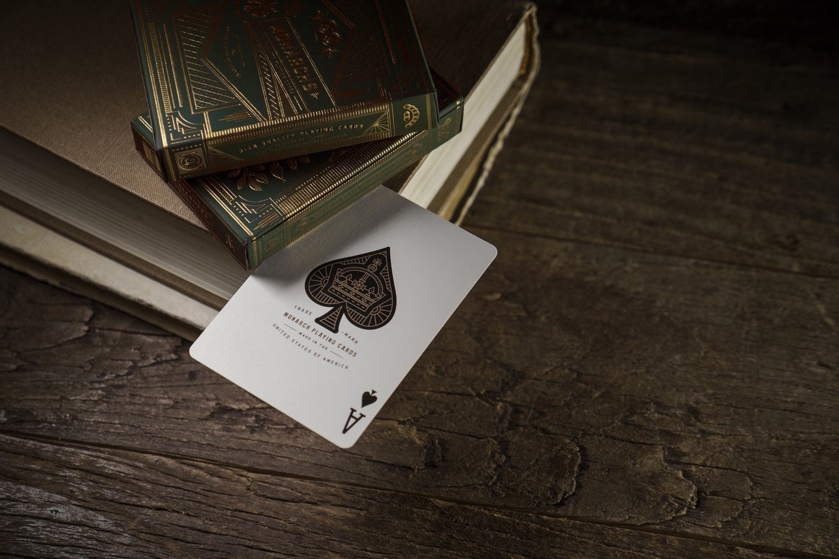 A detailed Ace of Spades playing card rests on dark wood, next to two ornate green and gold card decks on books