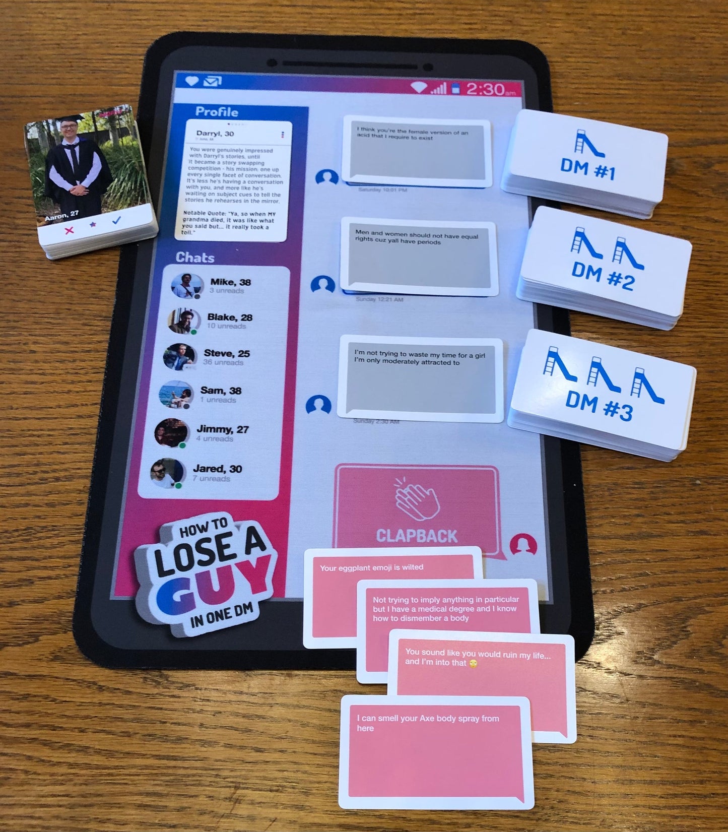 The How to Lose a Guy in One DM Card Game is laid out, featuring a phone-shaped game board with profiles and chat messages, alongside stacks of DM cards and pink reply cards