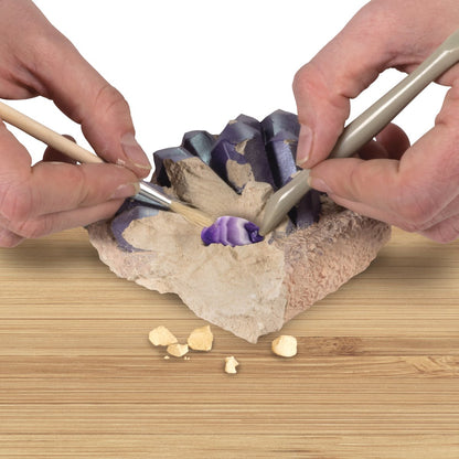 Hands excavating a purple striped crystal from a beige rock digging kit using a brush and scraper