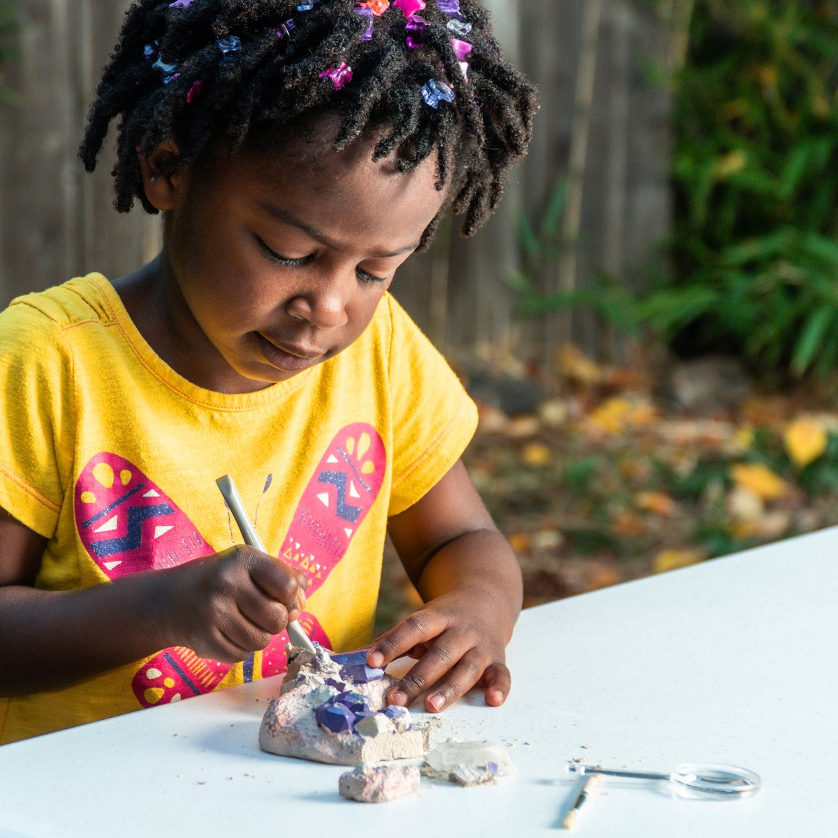 Child with beaded dreadlocks focused on digging purple gems out of an excavation kit on a white table outside