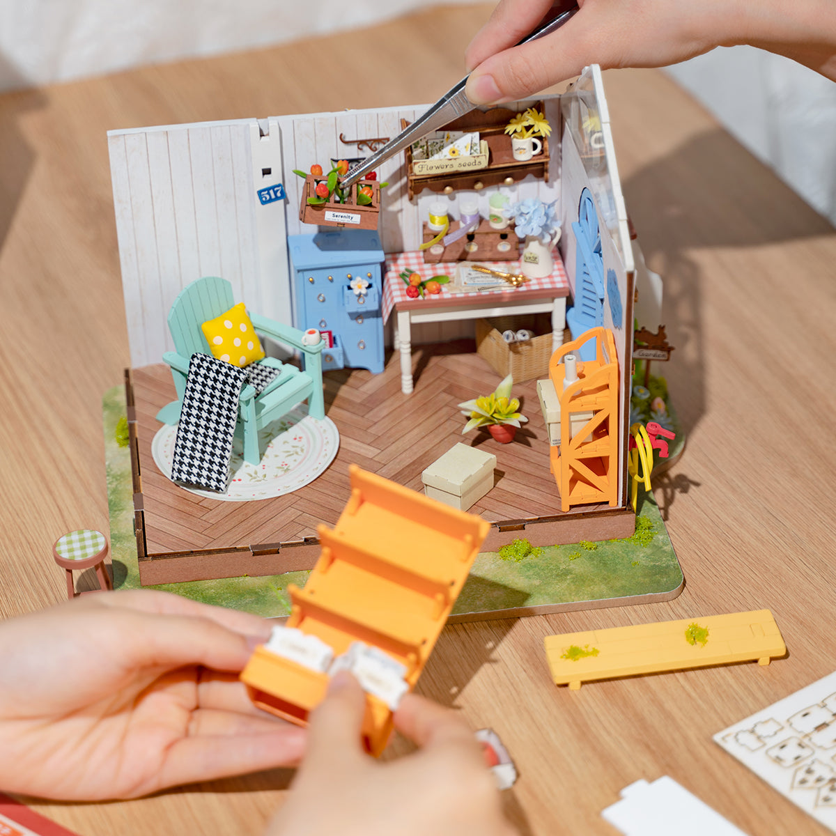 Hands assembling a miniature garden studio dollhouse kit; tweezers place tiny tulips into the scene