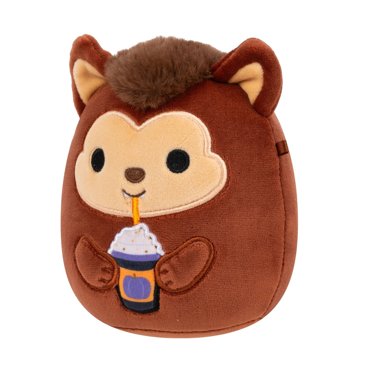 A brown Squishmallow plush toy with a light tan face, black eyes, and a fuzzy brown top, holding a purple and orange Halloween drink with whipped cream