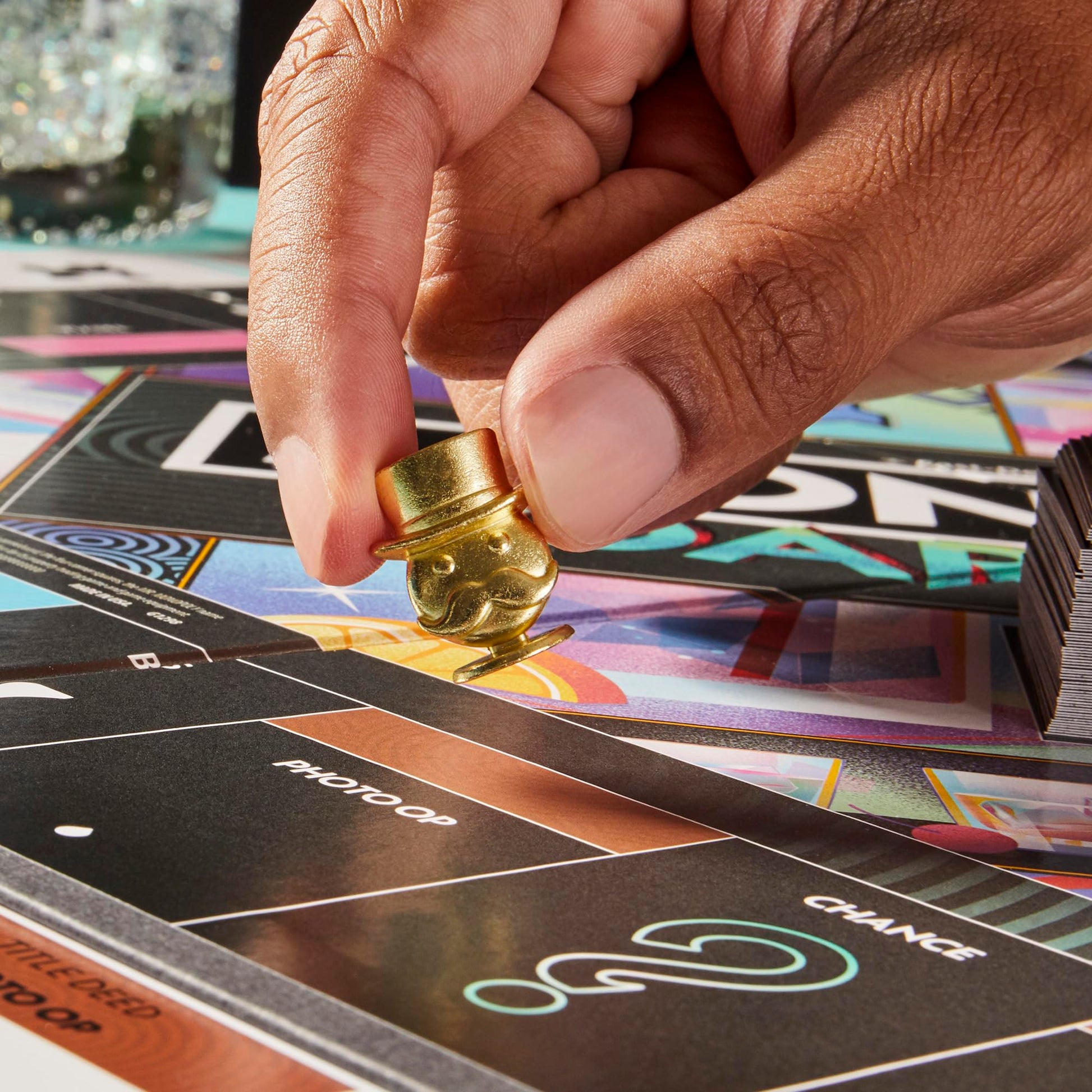 A hand places a gold top hat and mustache game piece on a colorful board with spaces like Photo Op and Chance, next to a stack of cards
