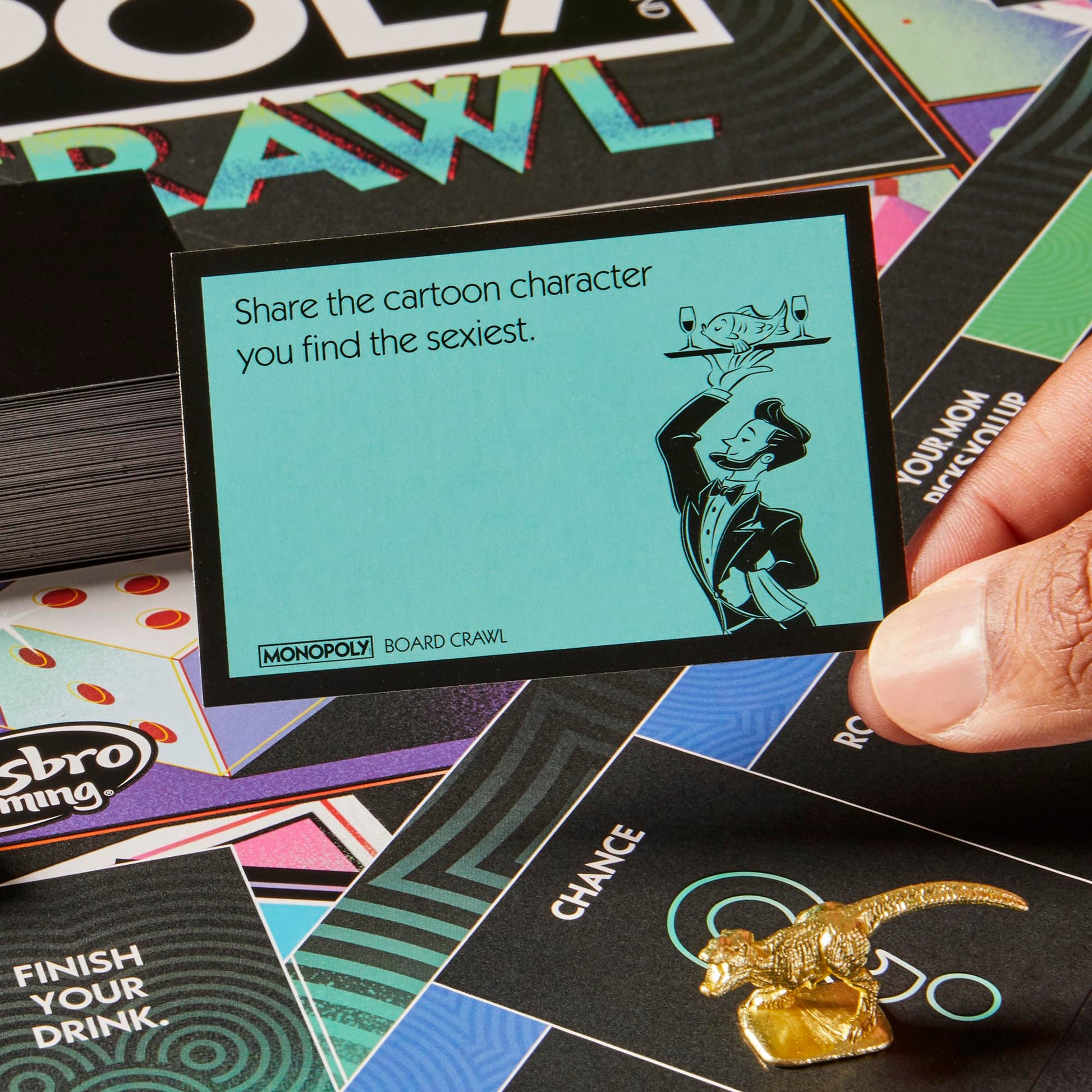 A hand holds a Monopoly Board Crawl card displaying a prompt about sexiest cartoon characters, with a butler illustration, alongside a golden dinosaur game piece on the board
