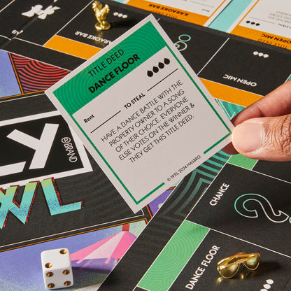 A hand holds a green Dance Floor title deed card over a Monopoly board, with a white die, gold sunglasses token, and a gold rooster token visible