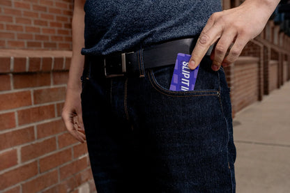 Hand slips a purple “SLIP IT IN” card into the front coin pocket of dark denim jeans