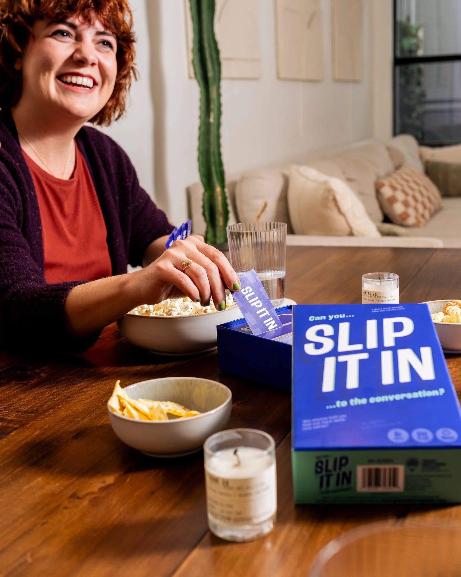 A smiling woman plays the card game "Slip It In," placing a card into the blue box next to snacks