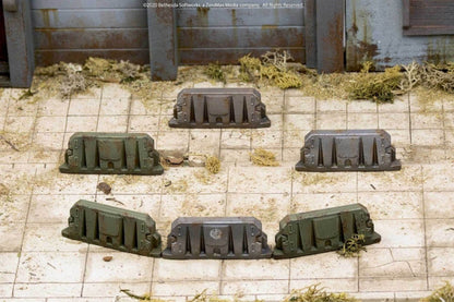 Six weathered green and grey miniature wargaming barriers arranged in a circle on a dirty tiled ground