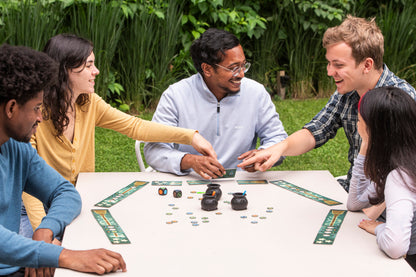 Five diverse friends happily playing a board game outdoors, features cards, dice, and small cauldron pieces are visible on the table