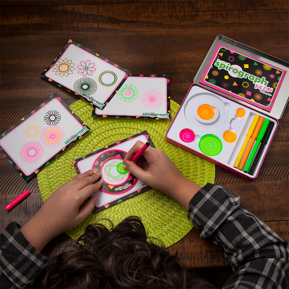 A person's hands use a Spirograph tool and pink pen to draw a bright green geometric pattern. Other colourful patterns and the kit are on the table.