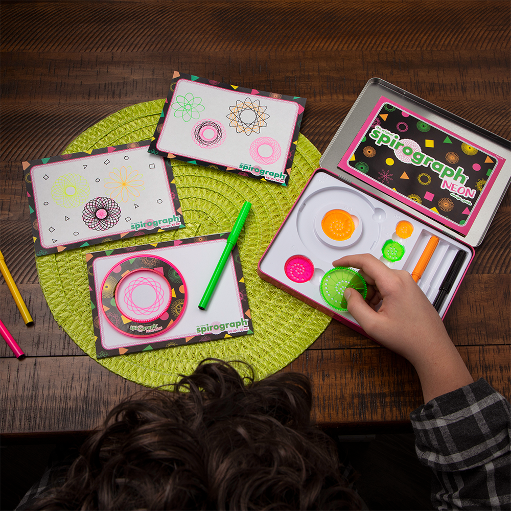 Person's hand holds a green gear from a Spirograph Neon kit. colourful geometric patterns, markers, and the kit are on a green placemat.