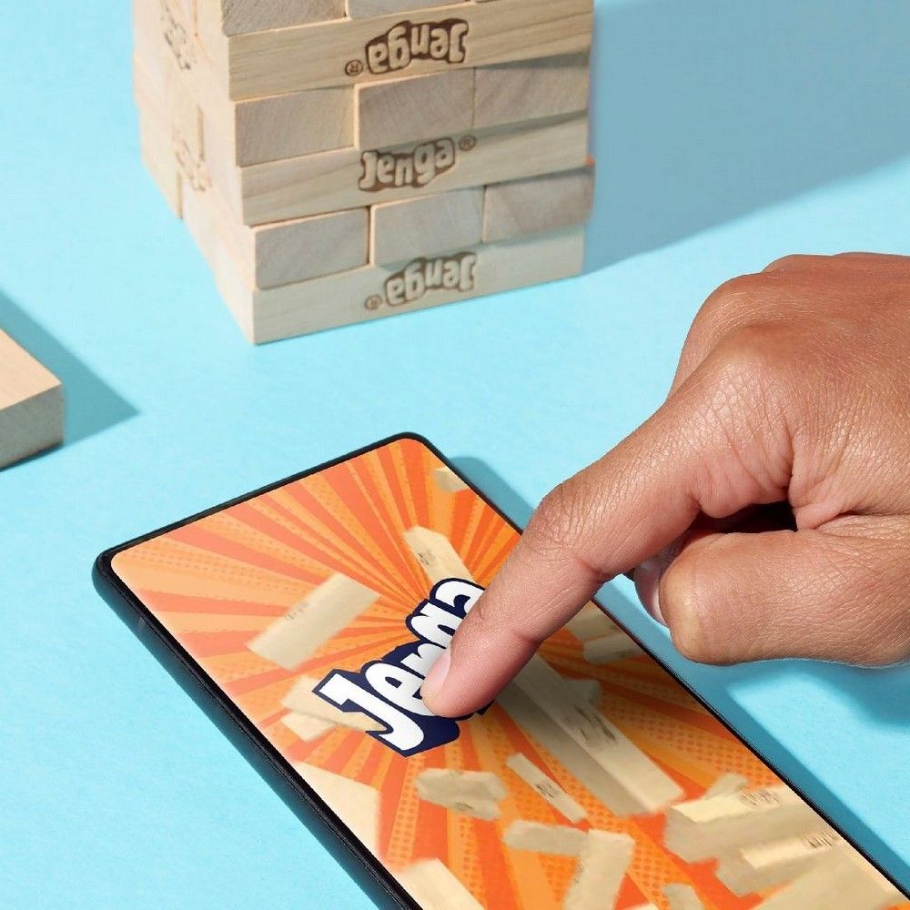 A hand taps a smartphone screen showing a Jenga game, with a physical Jenga block tower nearby