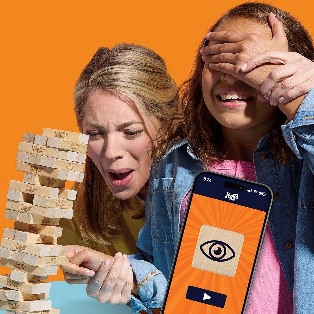 Woman plays Jenga, intently pulling a block. Girl covers eyes in suspense. Phone shows Jenga app with eye icon