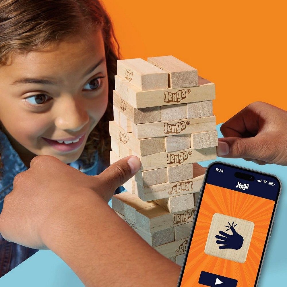 A smiling child carefully removes a block from a Jenga tower. A Jenga app is displayed on a smartphone beside it