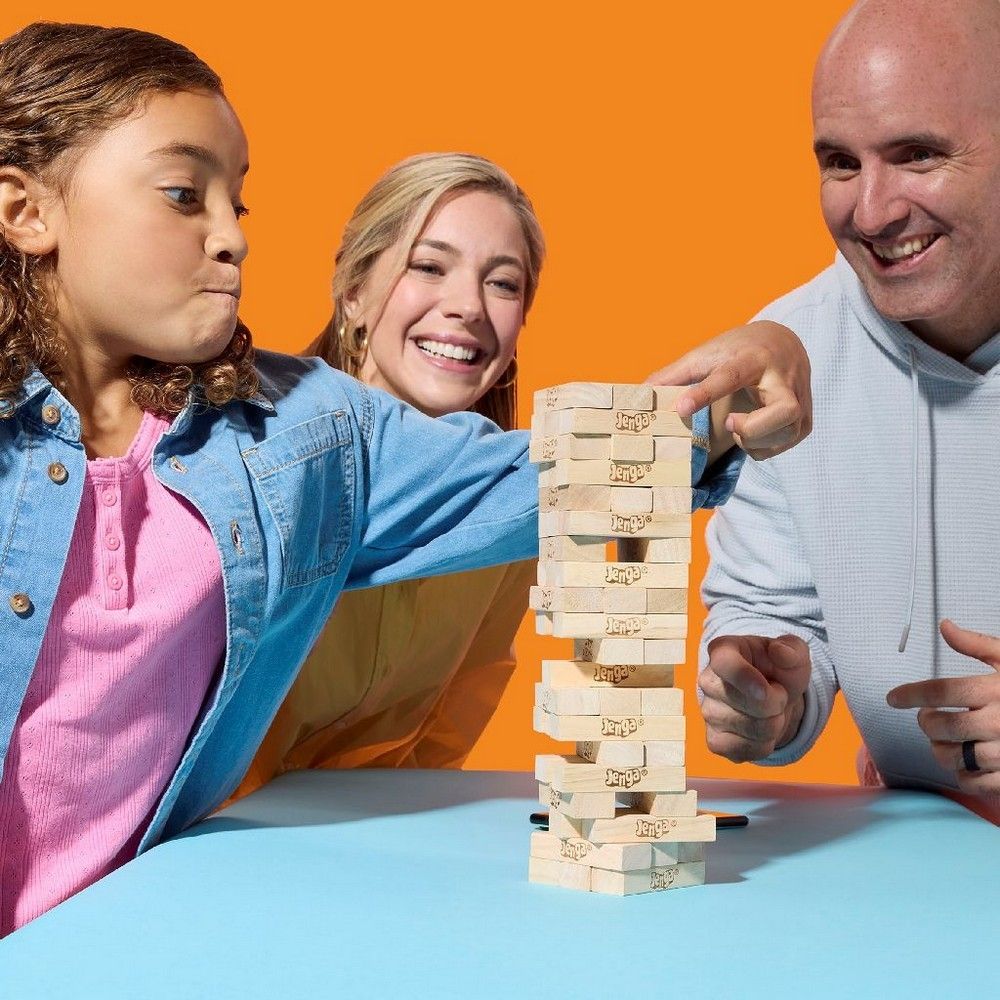 A man carefully pulls a Jenga block from a tall tower, watched by a girl making a focused face and a smiling woman