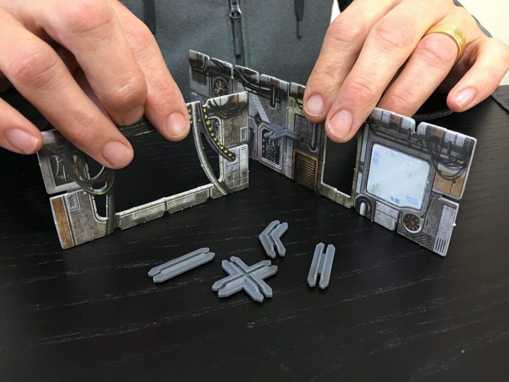 Hands assemble modular sci-fi game terrain walls with connectors on a dark table