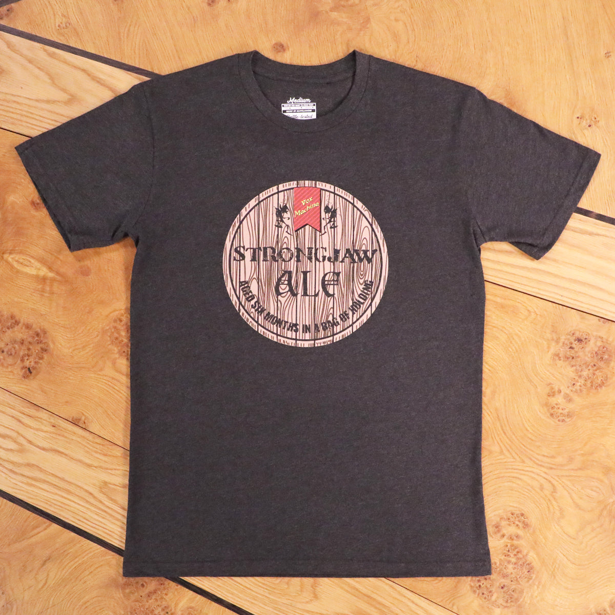 Dark grey t-shirt with a 'Strongjaw Ale' barrel label graphic including 'Vox Machina' logo, on wood