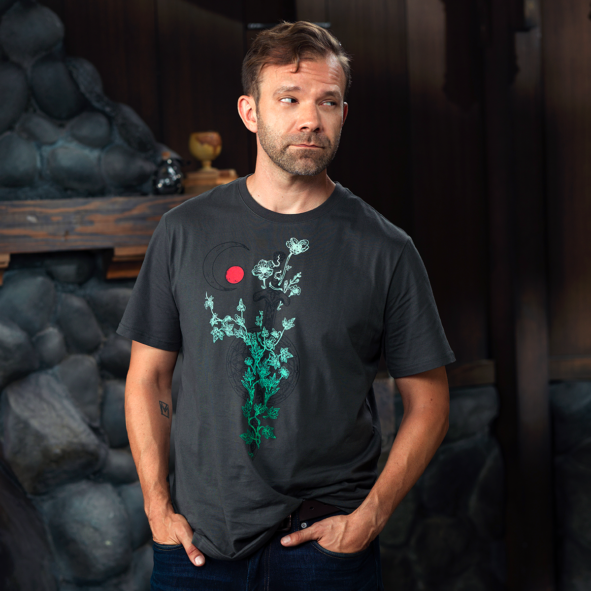 A man in a dark grey t-shirt with a green floral design and red circle stands with hands in pockets, looking left