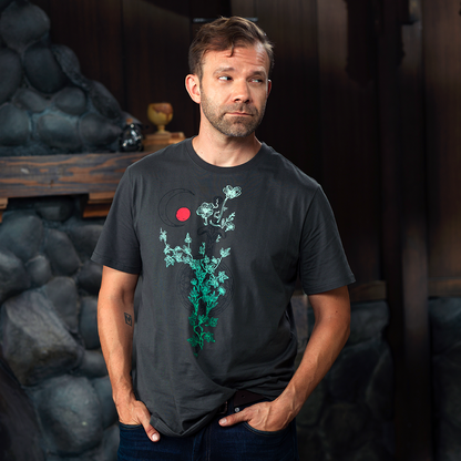 A man in a dark grey t-shirt with a green floral design and red circle stands with hands in pockets, looking left