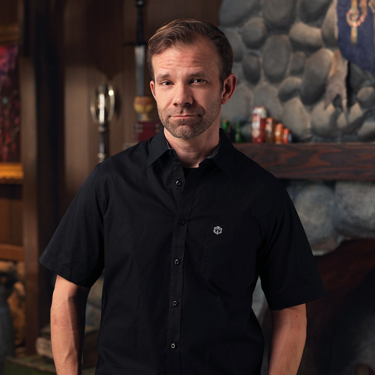 A man in a black shirt with a D20 logo looks at the camera. He stands before a stone fireplace with a sword and cans on the mantel