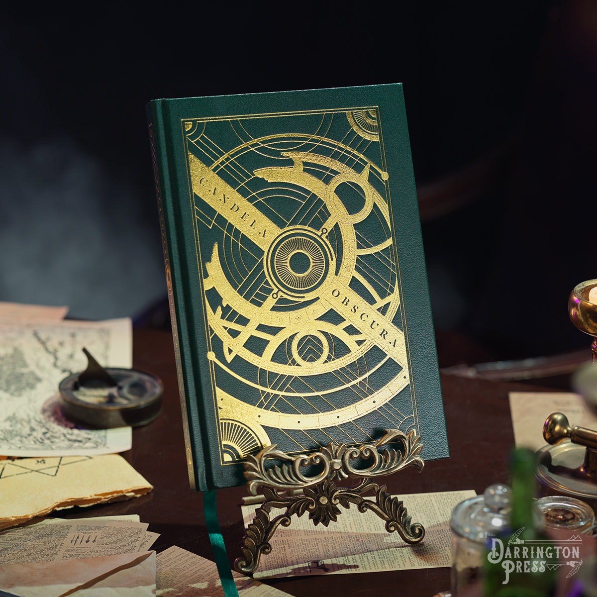 A dark green "Candela Obscura" core rulebook with a gold celestial design stands on an ornate display, amidst old maps and papers