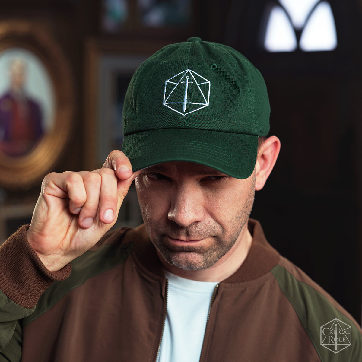 A man tips his dark green Critical Role Dad hat, featuring a white Critical Role d20 and sword logo, looking intently