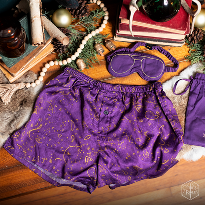 A purple Scanlan Shorthalt Boxers & Sleep Mask Set with shorts, an eye mask featuring gold glasses, and a matching bag, laid on a festive wood table