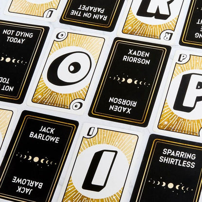 Black and gold playing cards, featuring text and symbols, including the name "Xaden Riorson," spread on a table