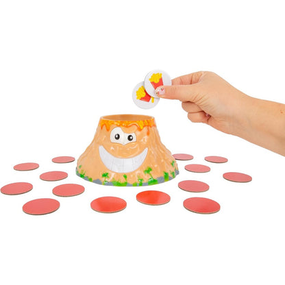 A hand holds two french fry tokens above the smiling lava-rimmed Volcanic Panic toy volcano surrounded by scattered red game pieces