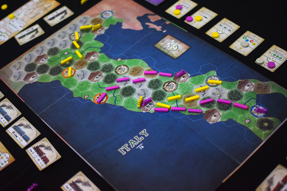 A board game map of Italy covered in yellow and pink train route pieces, buildings, and player cards