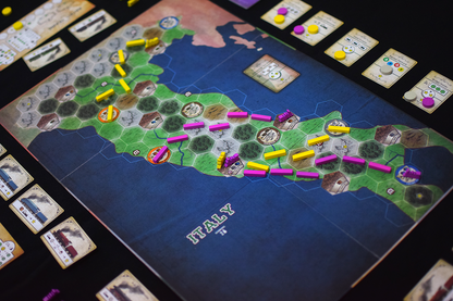 A board game map of Italy covered in yellow and pink train route pieces, buildings, and player cards