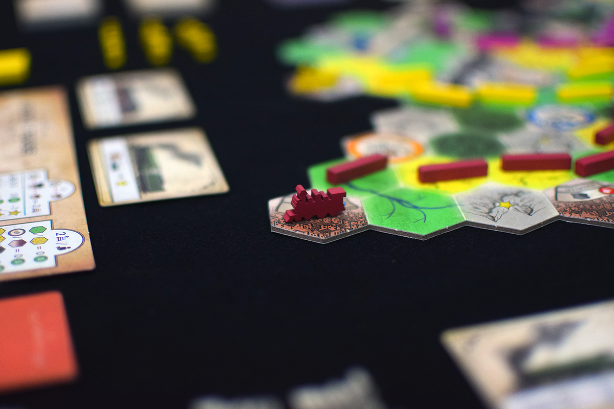 A red city piece sits on a hexagonal board game tile. Other game tiles, red train pieces, and cards are visible