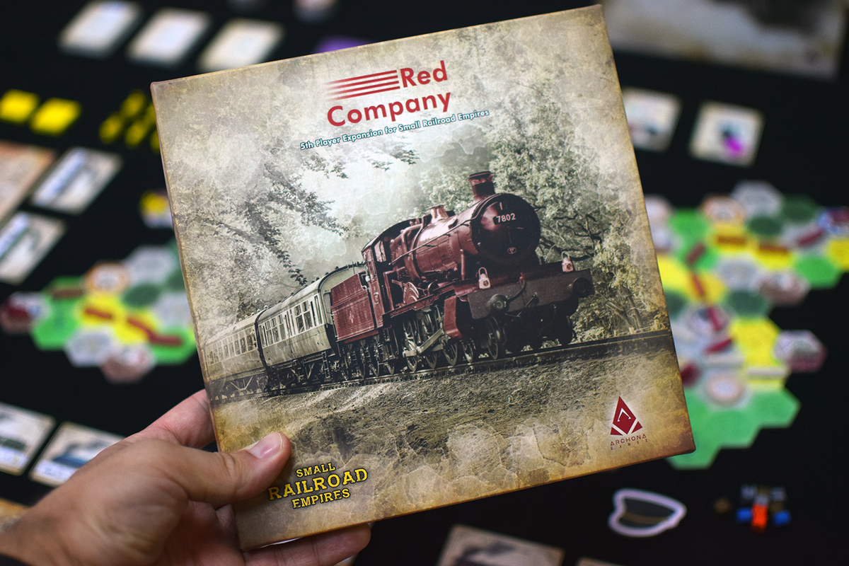 Hand holds "Small Railroad Empires: Red Company" board game expansion box showing a vintage red steam train. Blurred game board in background