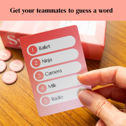 A hand holds a pink game card with a numbered list of words like Ballet, Ninja, Camera, Milk, and Radio, with game tokens and a box in the background