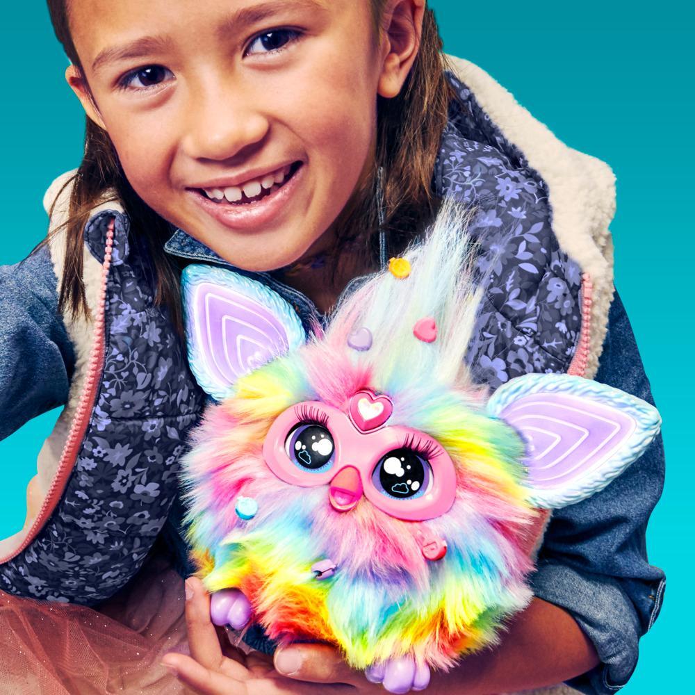 Smiling girl holds a fluffy, rainbow Furby toy with big eyes and glowing ears