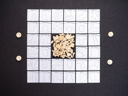 Tokyo Jutaku, a strategy board game, displays white grid-patterned cards with geometric building plots and city names, a pile of wooden shapes, and four tokens on a dark background