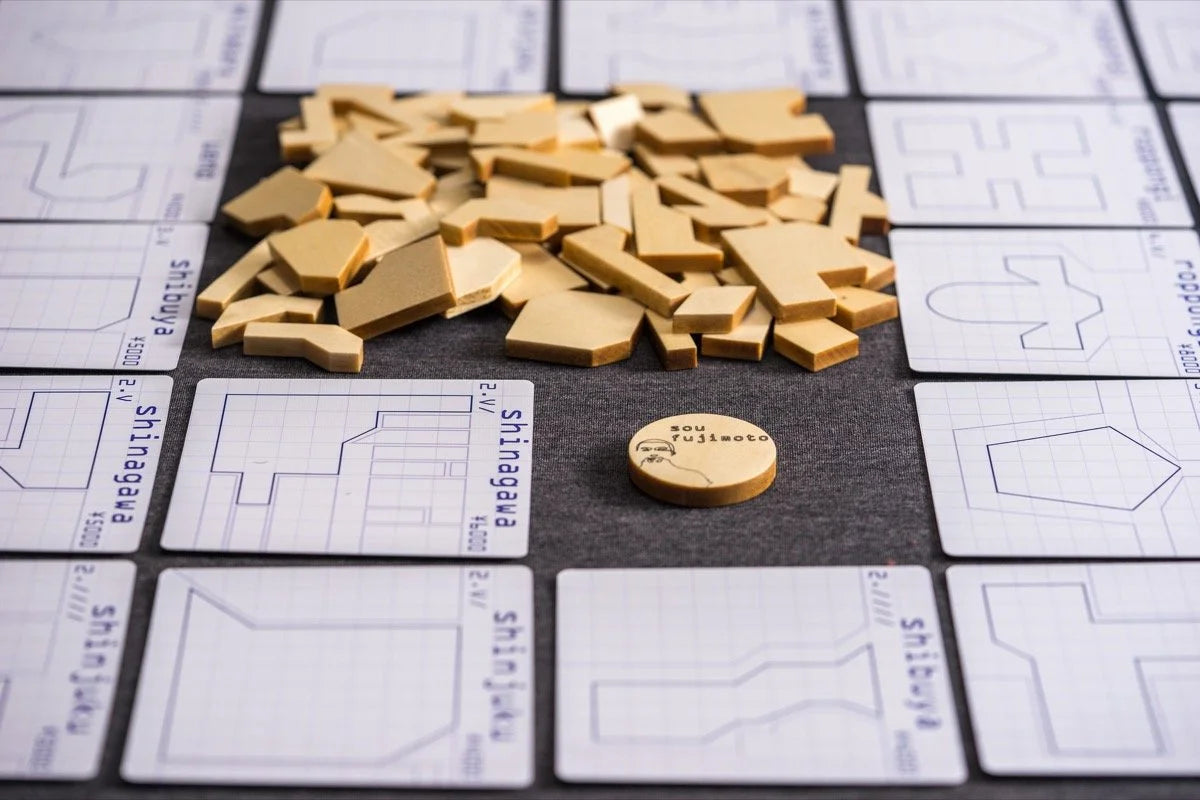A Tokyo Jutaku board game setup with numerous light brown irregularly shaped pieces, a round token, and grid cards showing architectural building outlines