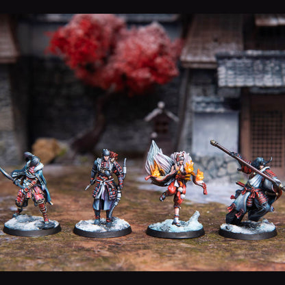 Four painted miniature Japanese warriors on snowy bases, posed in a blurred traditional village. One figure conjures fire, others carry weapons