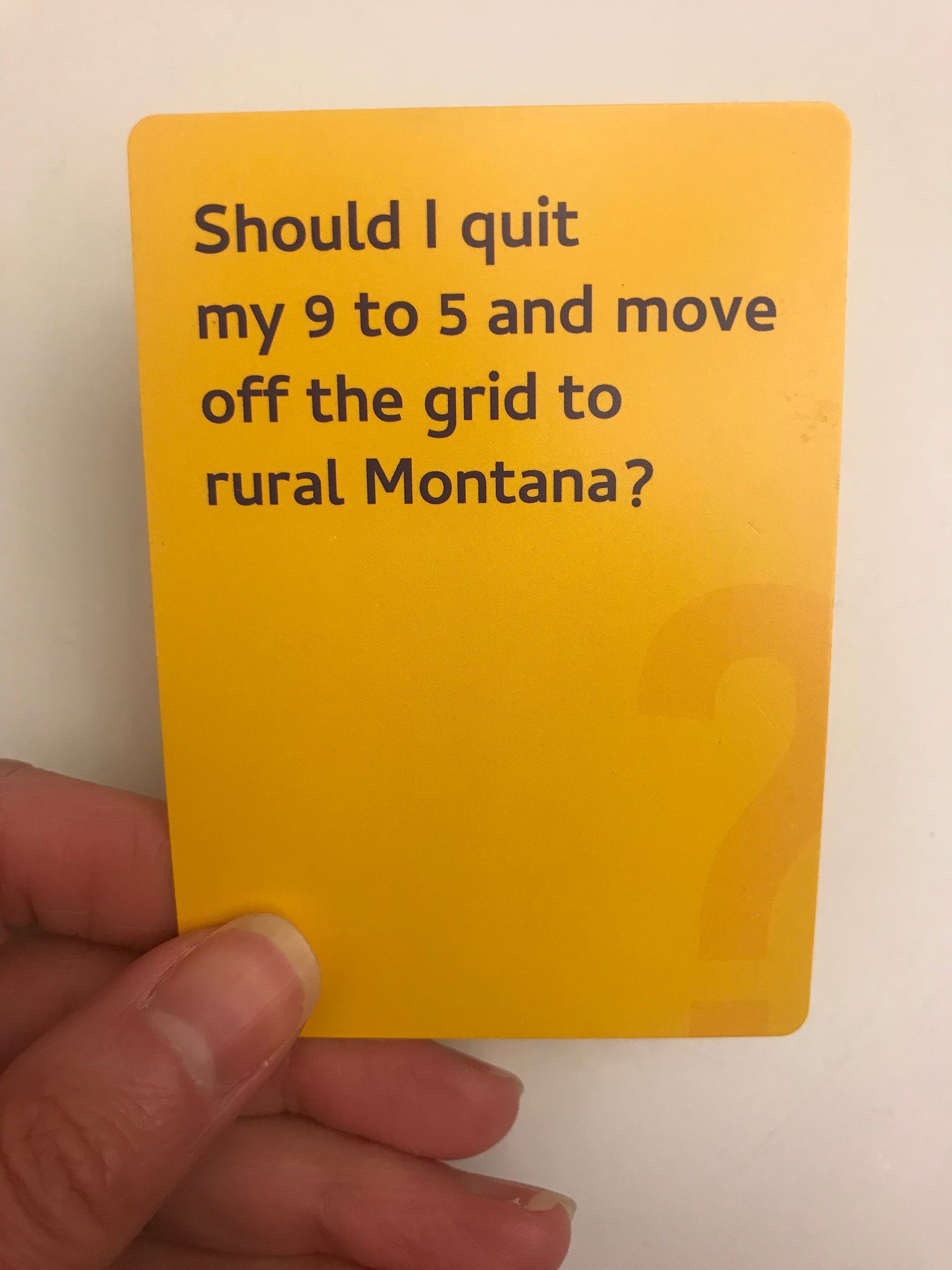 A hand holds a yellow card that asks Should I quit my 9 to 5 and move off the grid to rural Montana, with a faint question mark design