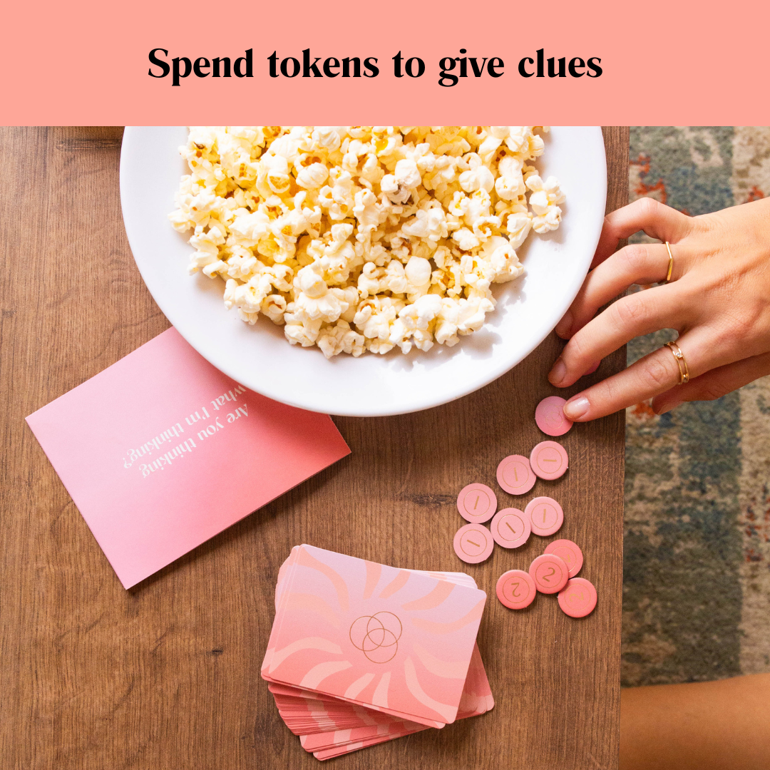 A tabletop game features pink cards, numbered tokens, popcorn, and a person's hand placing a token
