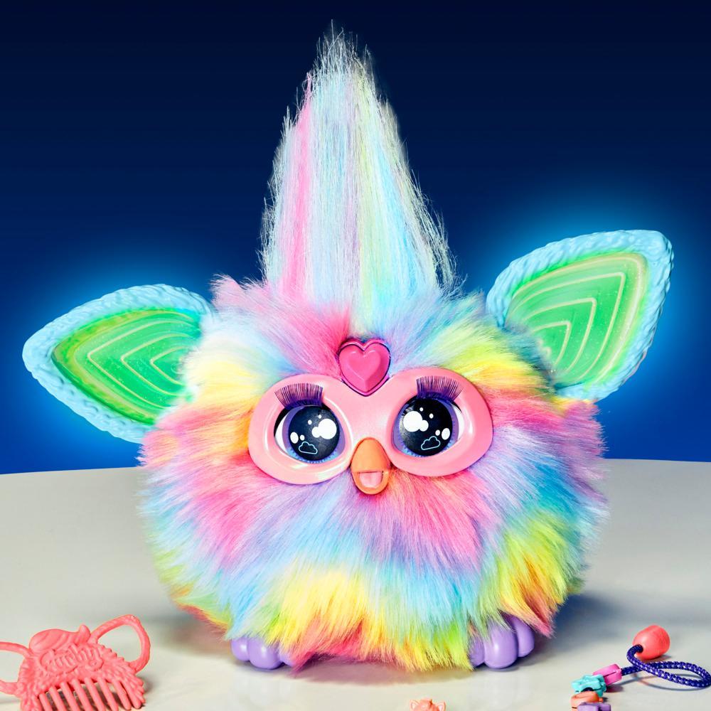 A fluffy rainbow Furby with big expressive eyes, long lashes, a pink heart, and glowing blue ears, next to a pink comb
