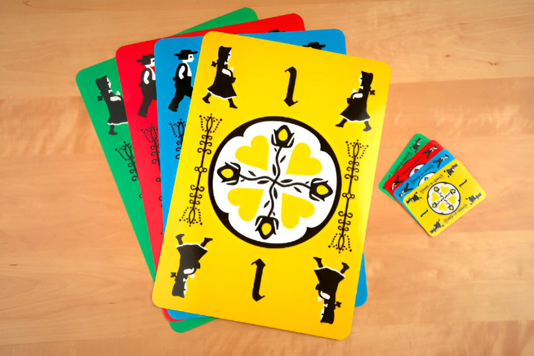 Colorful Dutch Blitz JUMBO! game cards fanned out on a light wooden table. A yellow card with &#39;1&#39;, character silhouettes, and a heart design is prominent