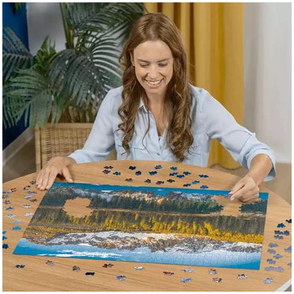 Woman smiling assembling a Ravensburger Lake Carezza 1000-piece jigsaw puzzle on a wooden table with many loose pieces surrounding it