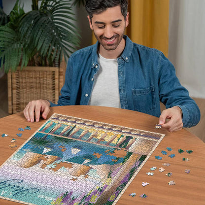Man smiling while assembling a Ravensburger Postcard from Capri Italy 1000-piece puzzle on a wooden table with scattered pieces nearby