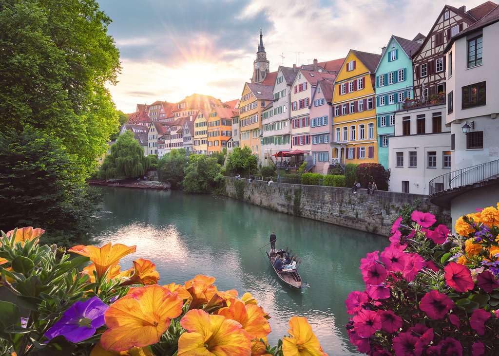 Picturesque 1000-piece puzzle artwork depicting Tübingen's colorful half-timbered houses lining the Neckar river at sunset, framed by bright orange and magenta flowers