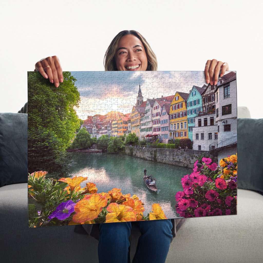 A smiling woman displays a completed 1000-piece Ravensburger puzzle featuring the colorful riverside houses of Tübingen Germany framed by bright flowers